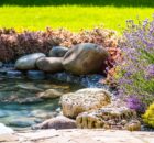 A beautifully manicured landscape with freshly cut grass, colorful flowers, and a stone-surrounded water feature.