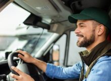 A smiling young man wearing a green baseball cap and driving a truck. He holds the wheel with both hands.