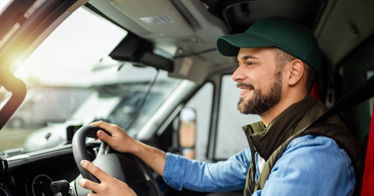 A smiling young man wearing a green baseball cap and driving a truck. He holds the wheel with both hands.