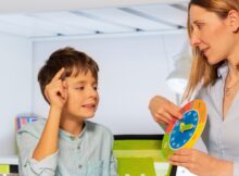 A woman sits in a chair holding a clock. A young boy sits next to her and is pointing at the clock.