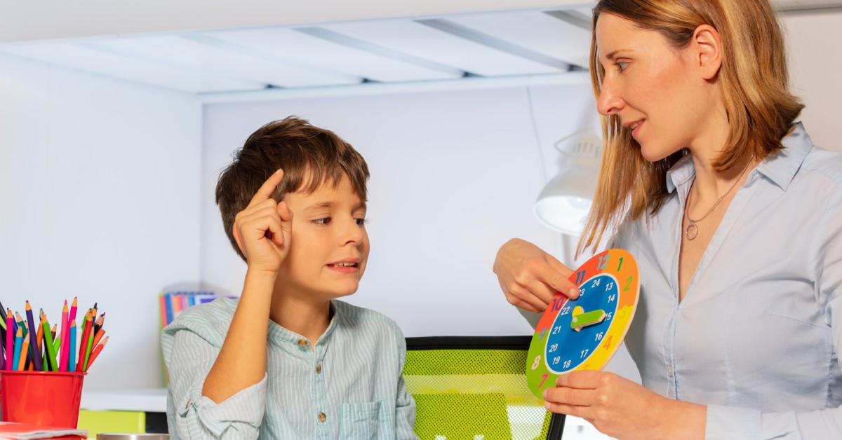 A woman sits in a chair holding a clock. A young boy sits next to her and is pointing at the clock.