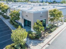 The outside of a commercial building strip is beige. There are several trees and bushes surrounding the businesses.