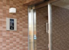 The exterior of a clean and secure residential building. There is an exterior light over an intercom panel outside the door.