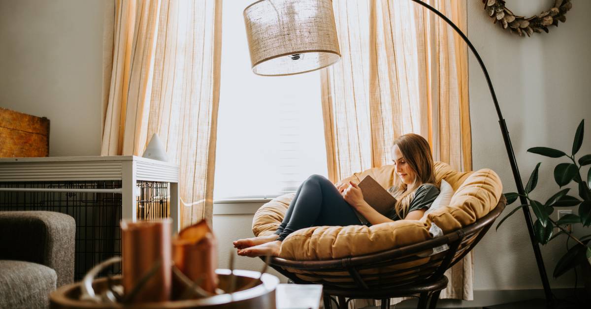 A young woman is sitting in a cozy chair next to a window reading a book. There's a tall lamp providing light.