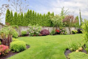 A fenced-in backyard with landscaping that includes lush flowerbeds throughout and a tight row of tall cypress trees.