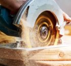 A man operates an electric saw over a large piece of wood. Sawdust falls off the back of the saw.