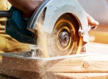 A man operates an electric saw over a large piece of wood. Sawdust falls off the back of the saw.