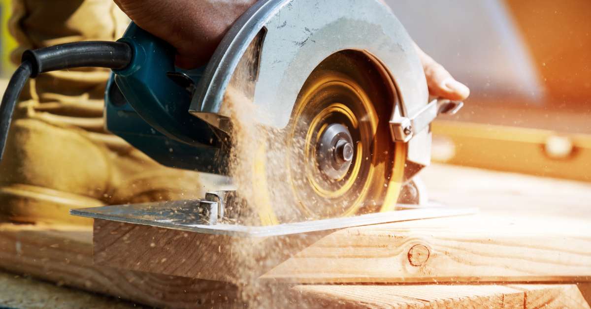 A man operates an electric saw over a large piece of wood. Sawdust falls off the back of the saw.