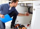 A repair technician is working on a homeowner's plumbing in a kitchen. The plumber holds a clipboard in their right hand.