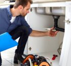 A repair technician is working on a homeowner's plumbing in a kitchen. The plumber holds a clipboard in their right hand.