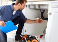 A repair technician is working on a homeowner's plumbing in a kitchen. The plumber holds a clipboard in their right hand.