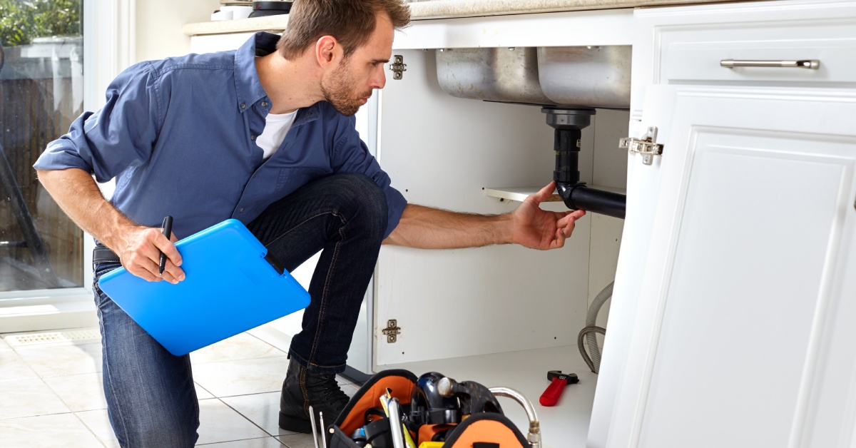 A repair technician is working on a homeowner's plumbing in a kitchen. The plumber holds a clipboard in their right hand.