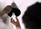 A woman holds the edge of an overhead pendant light fixture as she prepares to install a new LED light bulb.