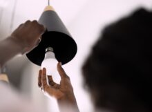 A woman holds the edge of an overhead pendant light fixture as she prepares to install a new LED light bulb.