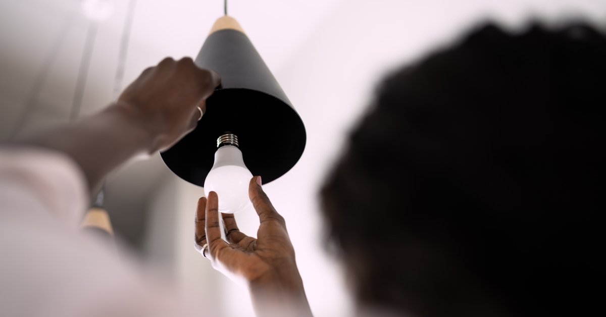 A woman holds the edge of an overhead pendant light fixture as she prepares to install a new LED light bulb.