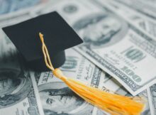 A pile of cash laying on a table with a collegiate graduation cap sitting on top of the cash. There is $100 bills.