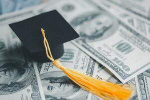 A pile of cash laying on a table with a collegiate graduation cap sitting on top of the cash. There is $100 bills.