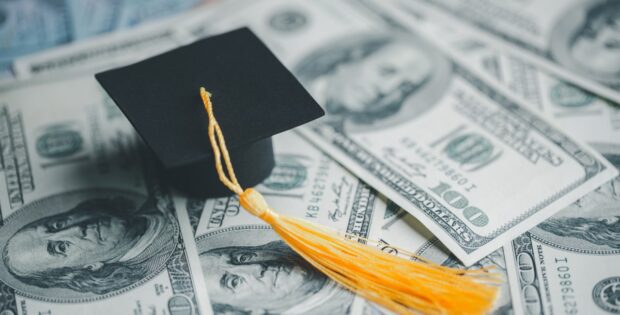 A pile of cash laying on a table with a collegiate graduation cap sitting on top of the cash. There is $100 bills.