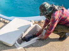 A man wearing a green camouflage hat is working on a pool pump system. The system is in a white compartment near the pool.