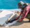 A man wearing a green camouflage hat is working on a pool pump system. The system is in a white compartment near the pool.