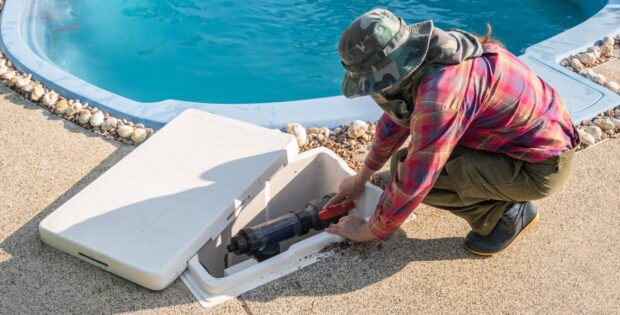 A man wearing a green camouflage hat is working on a pool pump system. The system is in a white compartment near the pool.