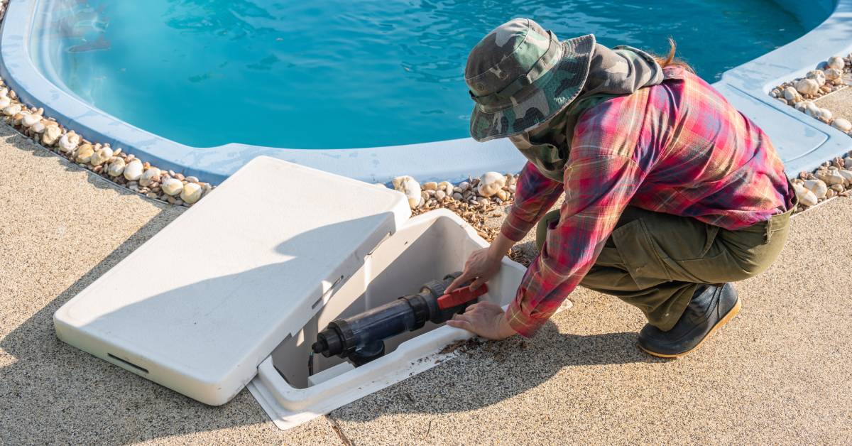 A man wearing a green camouflage hat is working on a pool pump system. The system is in a white compartment near the pool.